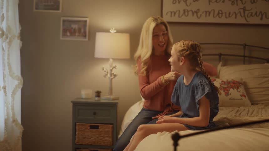 Mother braids daughters hair as they talk and laugh while sitting on bed