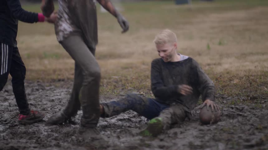 Child tackles another child in the mud while playing ball and a third child helps helps the first up