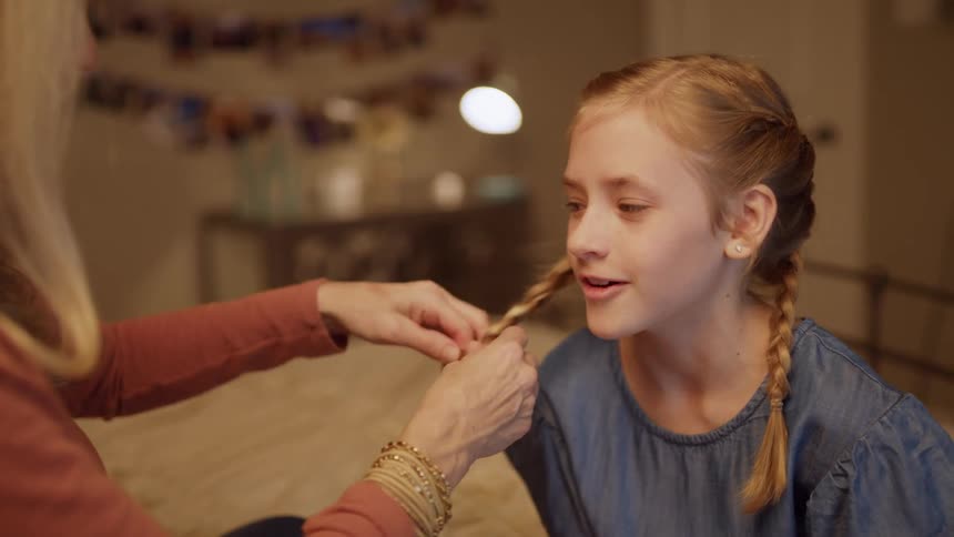 A young girl sits on a bed smiling and talking with a woman who braids her hair