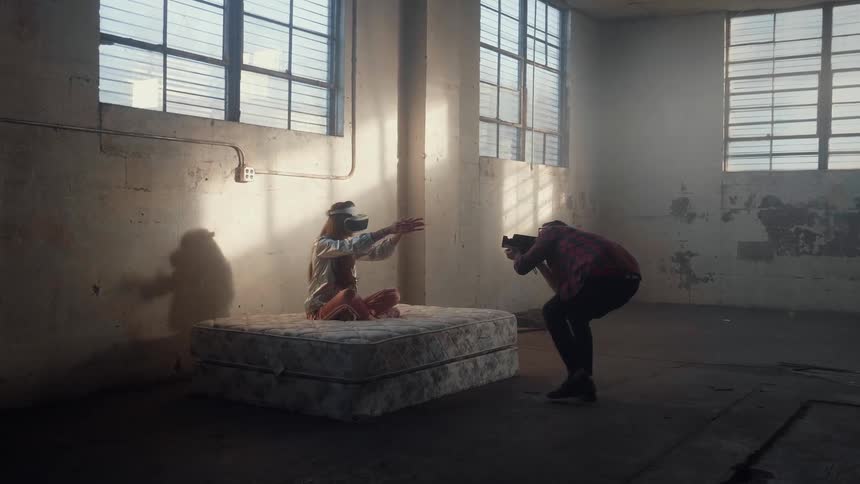 Woman sitting on mattresses in a building wearing vr gear as a man films her