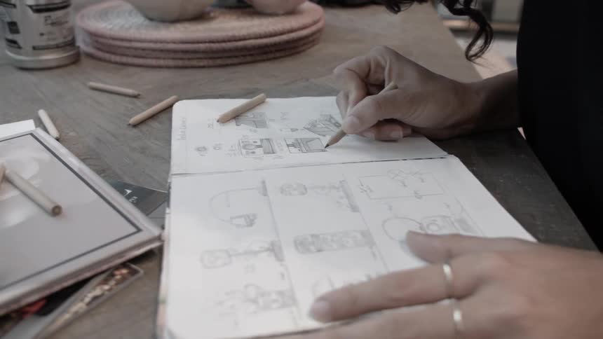 A woman sitting at a desk in a studio works on a pencil sketch in a book