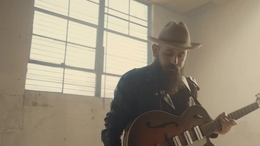 A man stands in an empty room with sun shining through the windows playing guitar