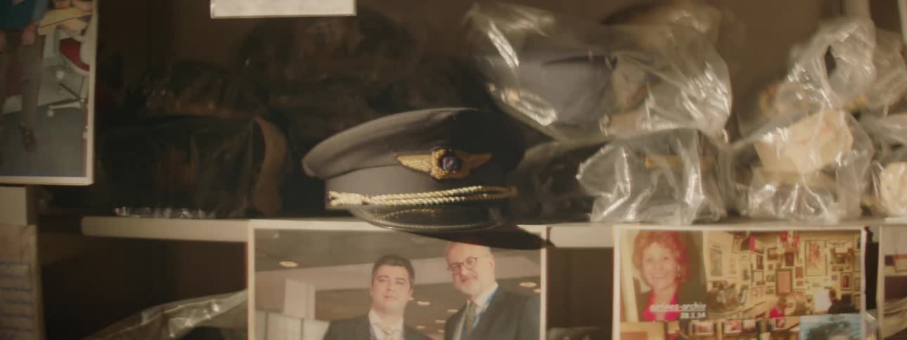 Hat on a shelf with many other things wrapped in plastic and pictures below