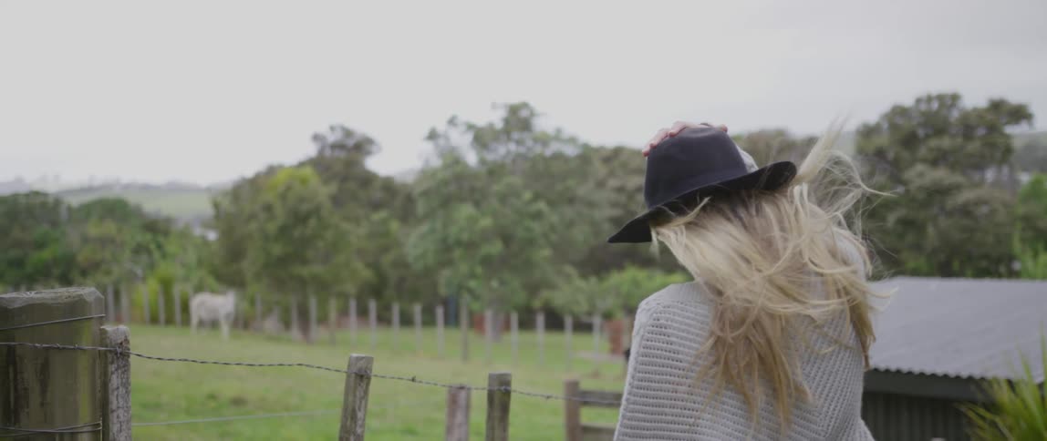 A white woman walks on a farm while holding her hat to her head in the wind