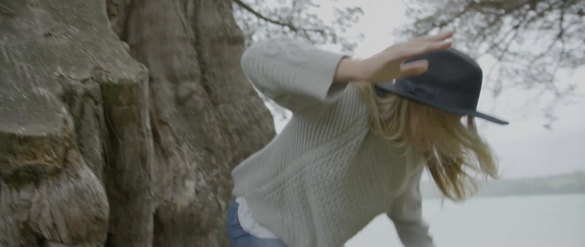 A blonde white woman wearing a hat jumps off rocks by a beach