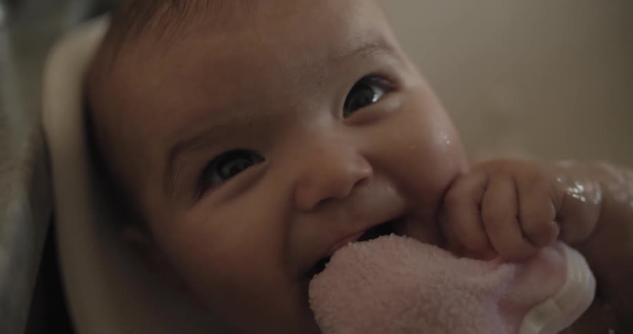 A baby in a bathtub sucks on a washcloth and smiles