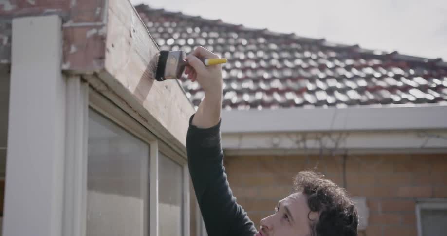 A man is dipping a paint brush in a can of paint and is painting part of his roof