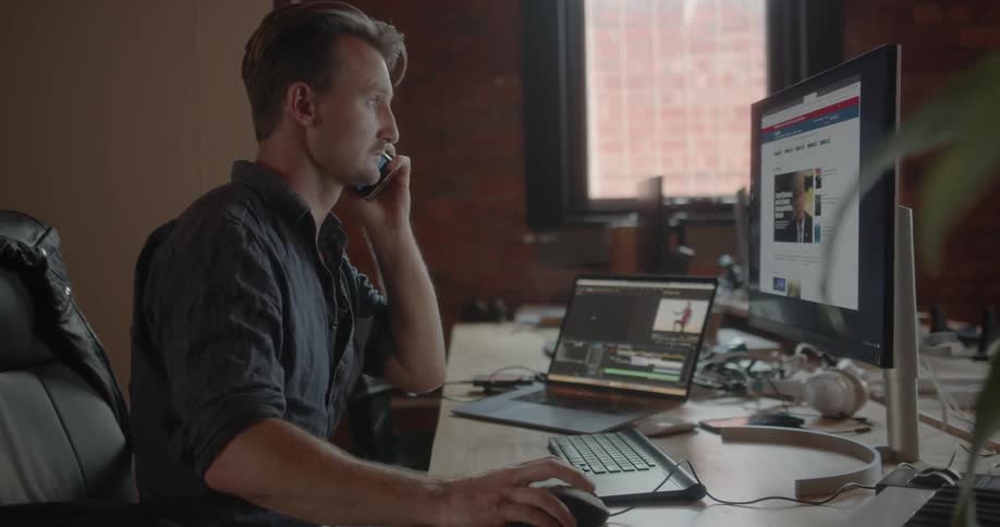 A man sitting at a desk in an office answers the phone while he works on his computer