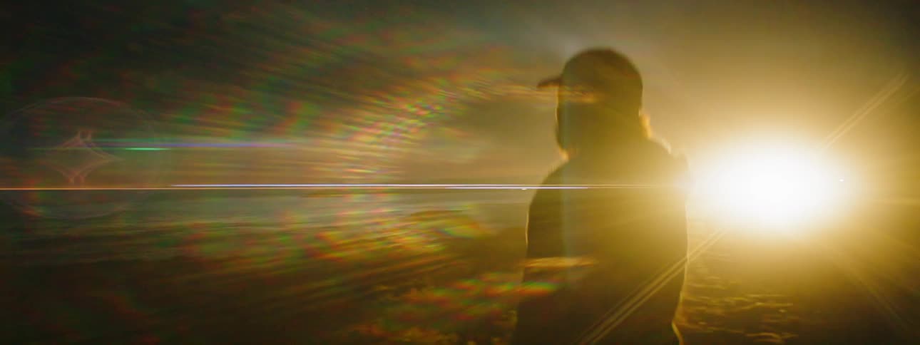 A person is standing on the shore at dawn with a light flashing in the distance