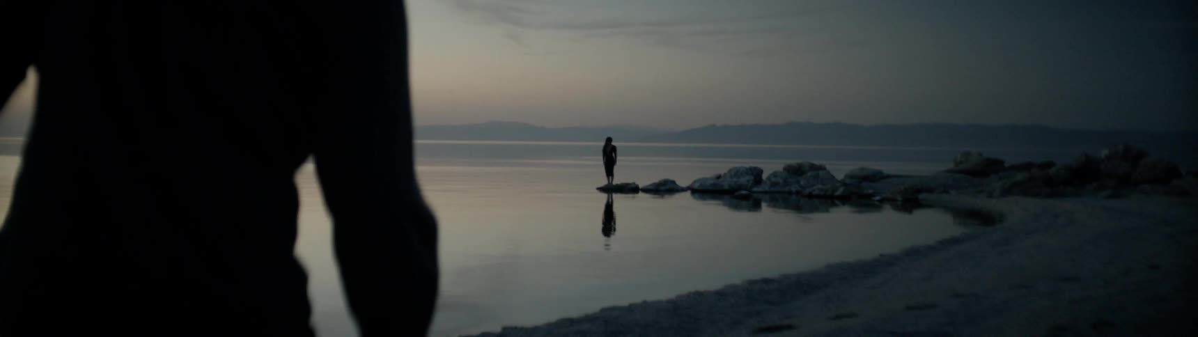 Person stands on a rock out into the water while another person walks on the beach at sunset