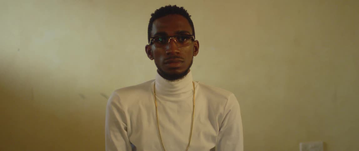 A man wearing glasses and a gold chain turns to stare forward