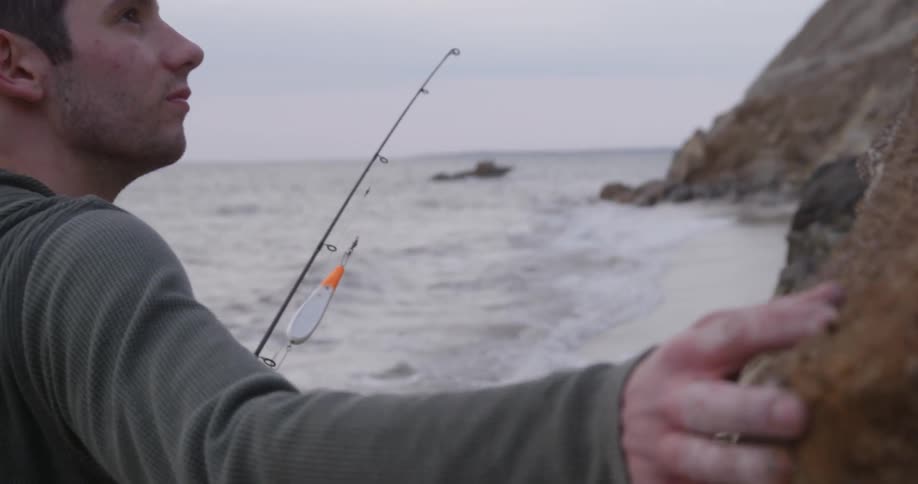 Man holding fishing pole traces hand across a cliff by the shore
