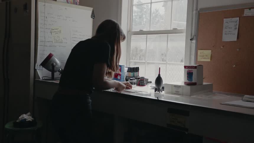 A woman leans on a counter near a window and looks at what she's holding