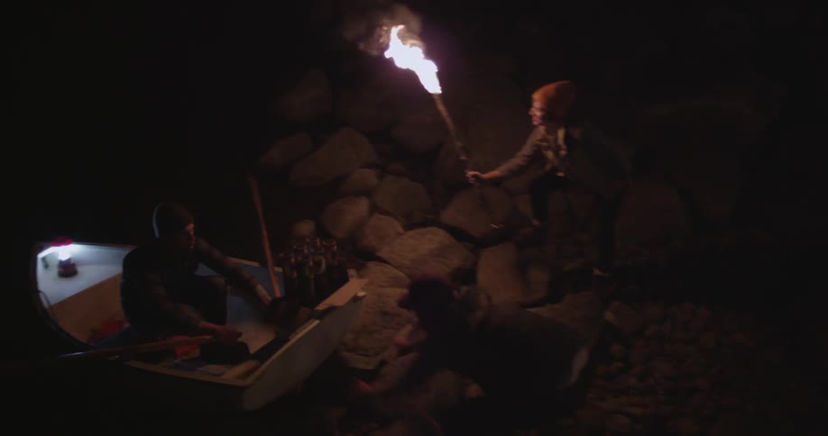 A man and a woman carrying a torch walk from a small campfire to push a small rowboat into the water while the occupant rows away into the darkness
