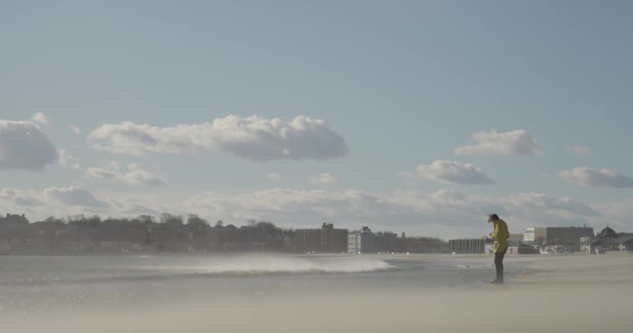 A man in a yellow jacket taking pictures from the shoreline as the wind whips around him
