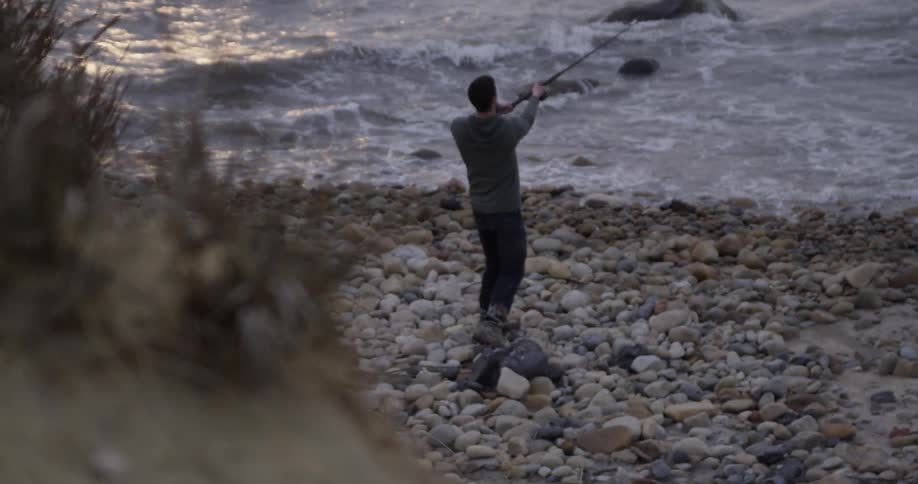 A man casts a fishing line on a rocky beach