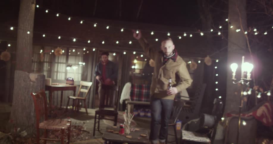 People running out of a house, carrying bottles of beer in their hands while they run under lights strung between trees and wave each other on