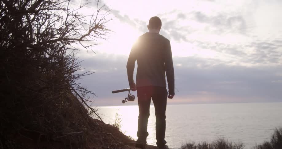 A man holding a fishing pole walks down a hill toward the beach in the distance