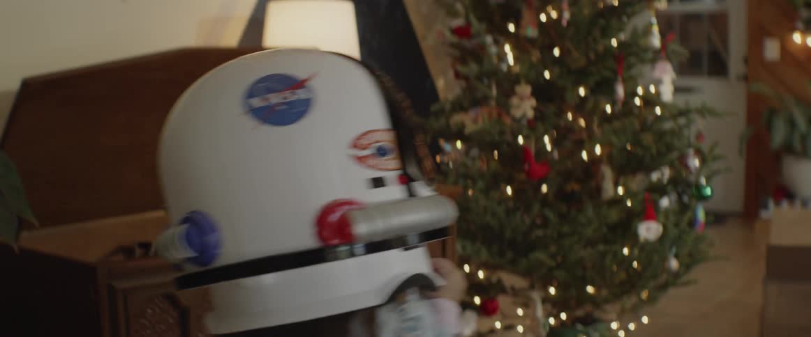 Small child wearing astronaut helmet walks past christmas tree towards mom