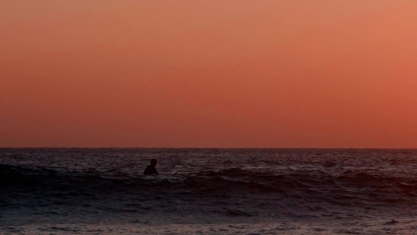 A surfer sits on his board and rides over a wave at sunset