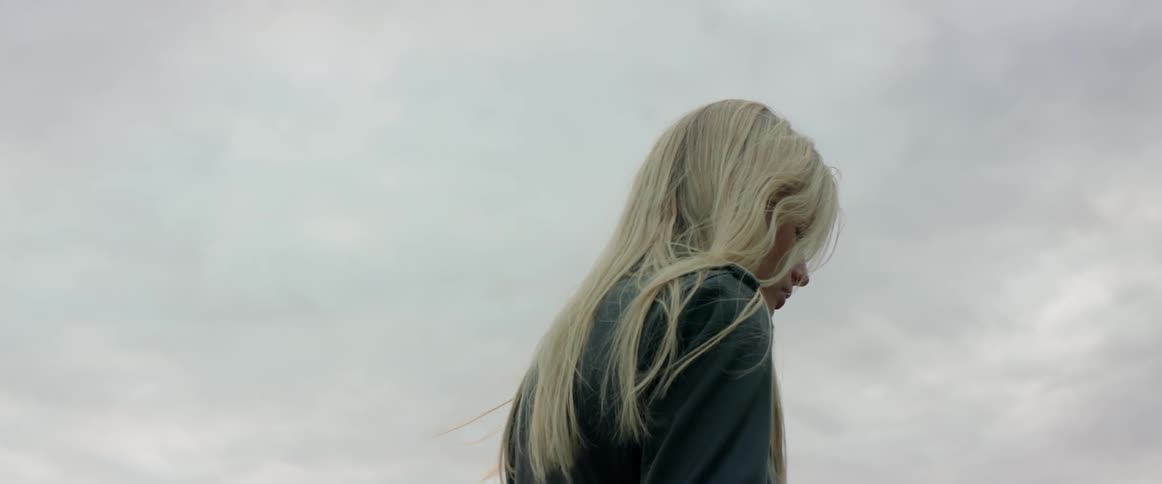 A sad blonde woman looks down thoughtfully while standing outdoors on cloudy day.