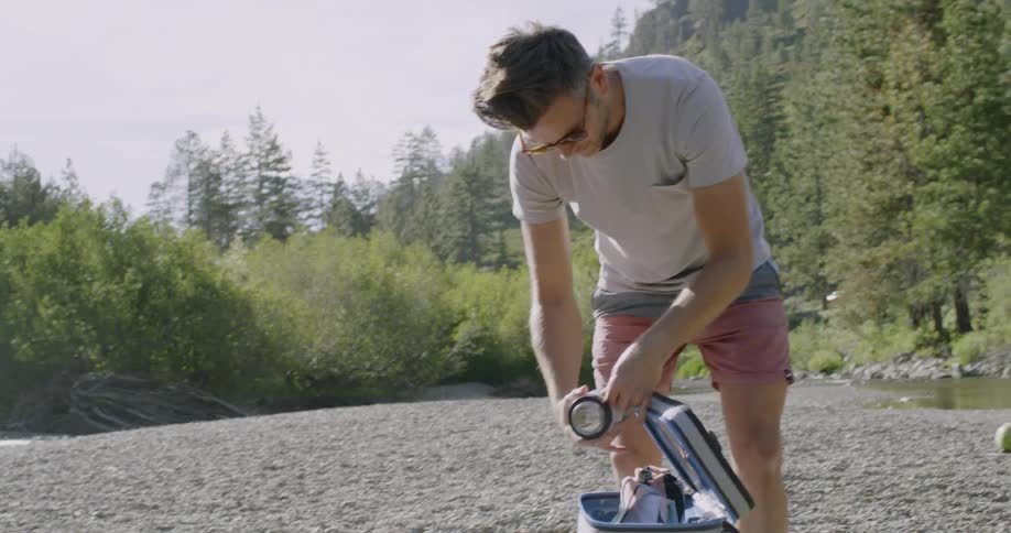 A man is unwrapping a container he is removing from a cooler in a natural setting in daylight