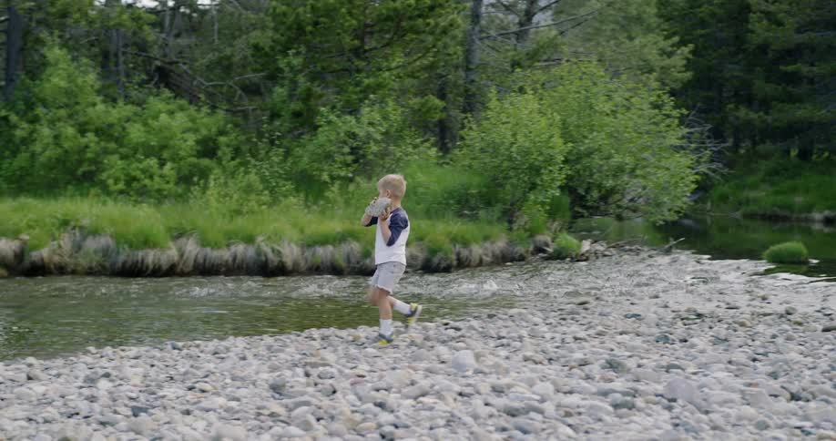 Little boy carries big rock to the river shore and throws it into the water