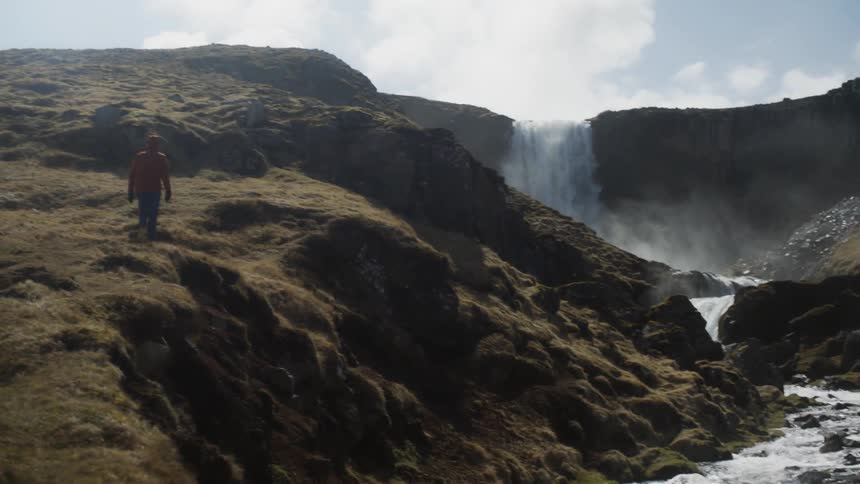 A man walks along a ridge towards a waterfall