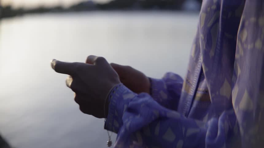 An anxious person checks her mobile phone while standing near a calm lake.