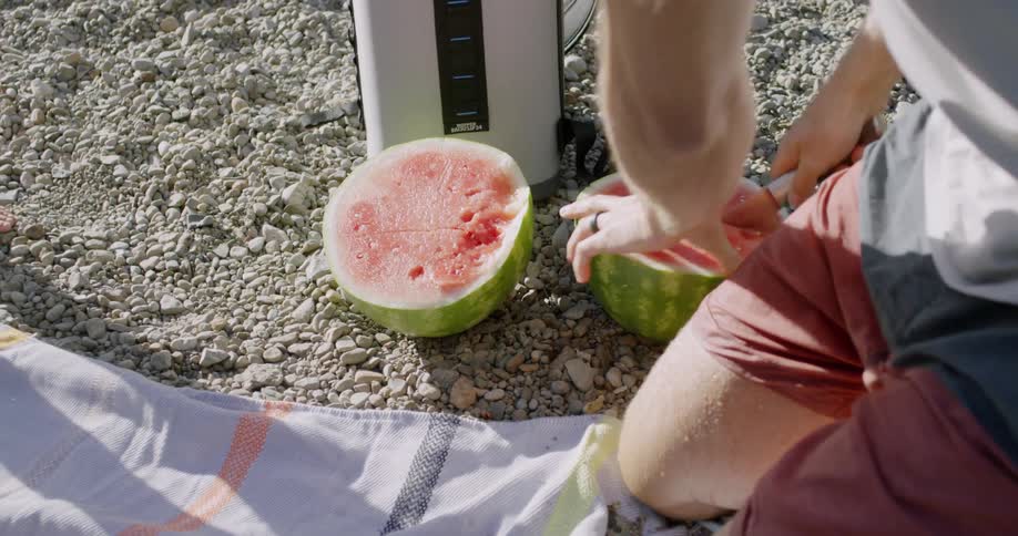A man cuts a watermelon while kneeling on a blanket outside