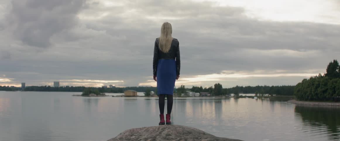 A lonely female traveler stands on a stone shoreline overlooking a calm lake.