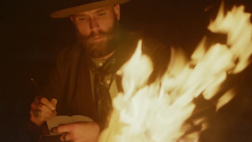 Bearded man writing in a book while next to a large campfire at night