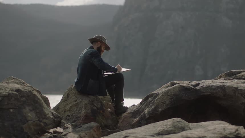 Man sits on a large boulder near water drawing a picture