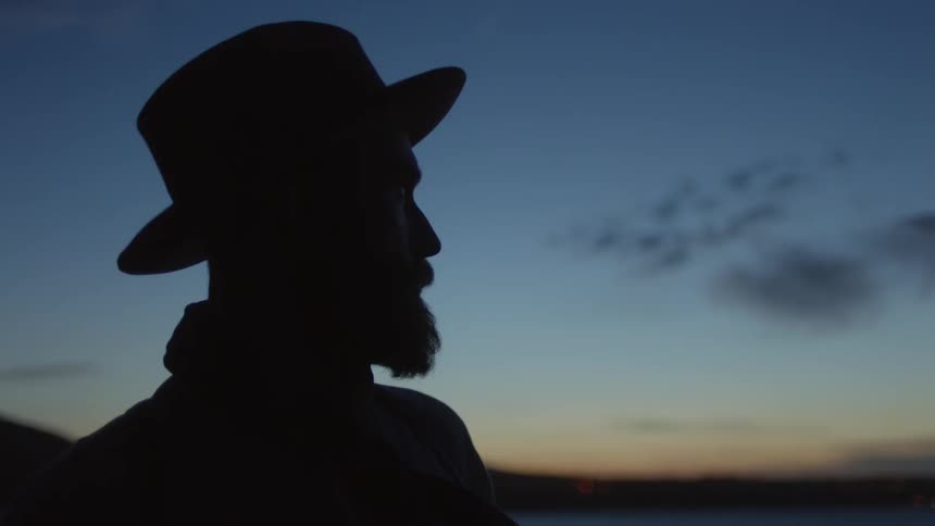 A man in a hat stands outside at dusk