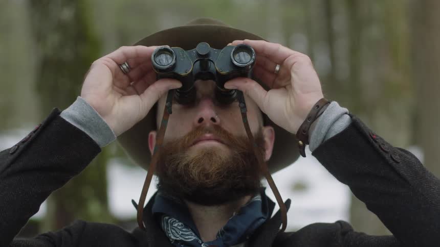 Man wearing hat is outside looking through binoculars