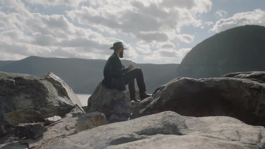 A man is sitting on a boulder above a lake writing in a notebook during the day