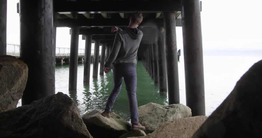 A young man standing on boulders under a pier while skipping stones inn the water