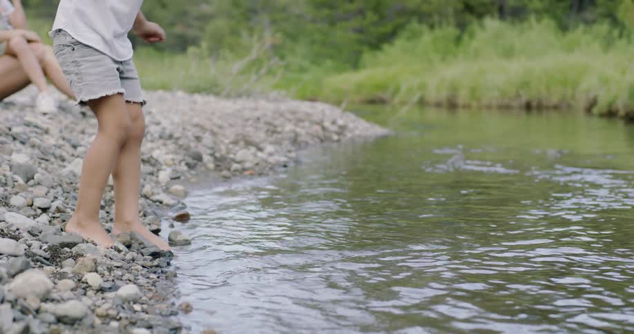A boy is edging off a riverbank and towards the water in daylight