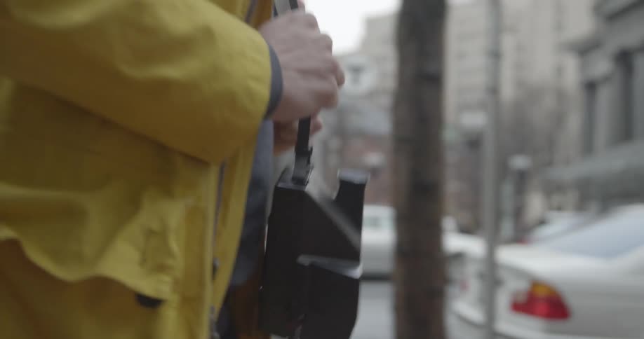 On a wet day a woman wearing a raincoat walks along a downtown sidewalk with polaroid camera dangling from a strap around her neck