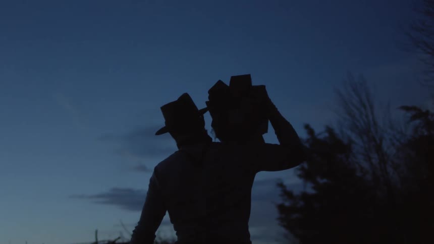 A man is carrying a stack of wood on his shoulder as he walks outside at dusk