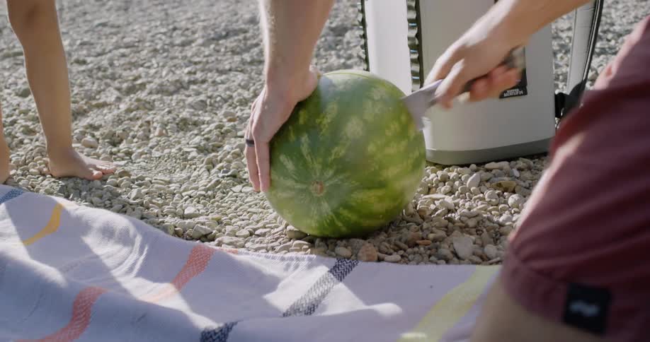 A man is using a knife to cut open a watermelon outside on a beach in sunlight