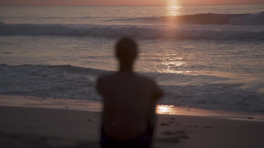 Man sits on beach and watches tide as sun sets