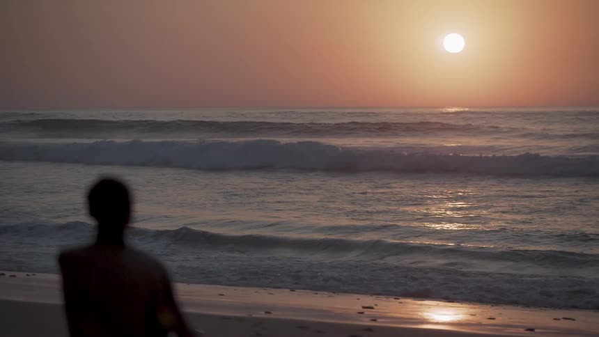 Man sits on beach observing waves rolling in as sun sets
