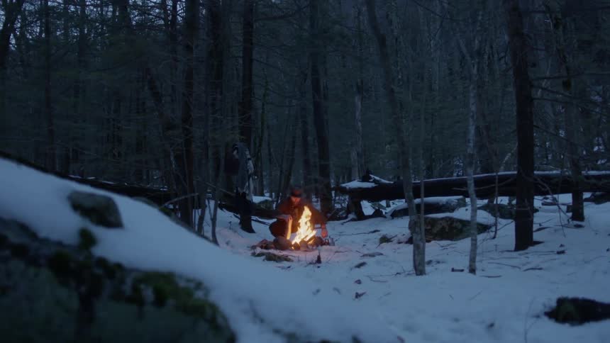 Man adds wood to campfire surrounded by snow at dusk