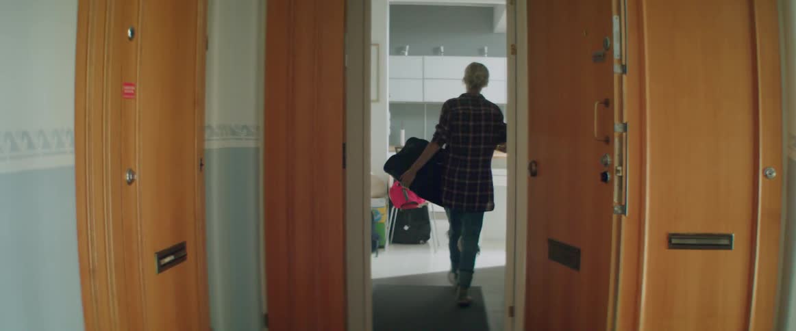 Rear view of a woman walking into an apartment while carrying large bags.