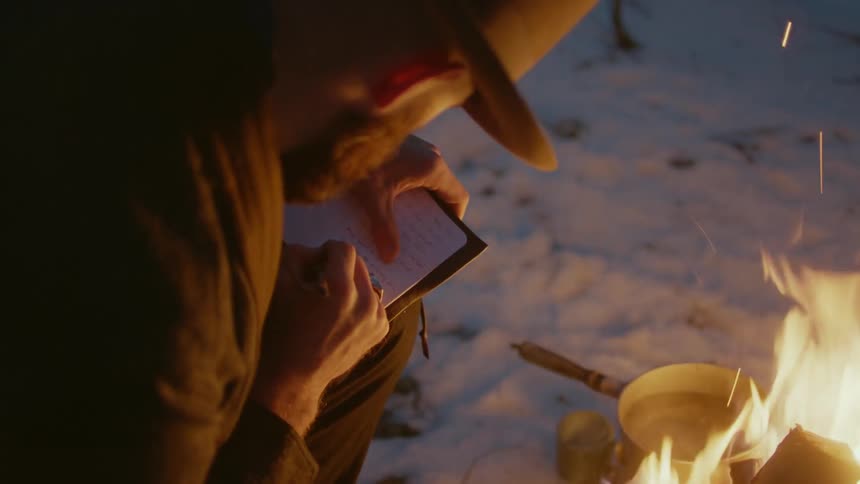 Man writes in notebook next to campfire surrounded by snow