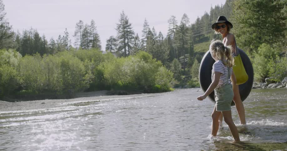 A mom carrying a tube holds her daughter's hand as they wade into the river