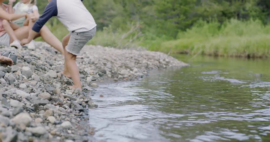 Little boy walks out of river water and up rocky shore where his family sit