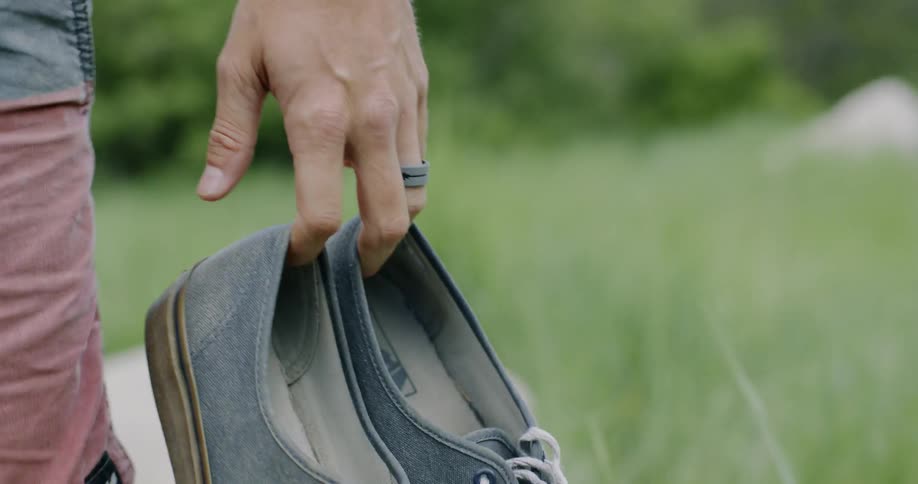 A man is carrying his shoes in his hands as he walks in a natural setting in daylight