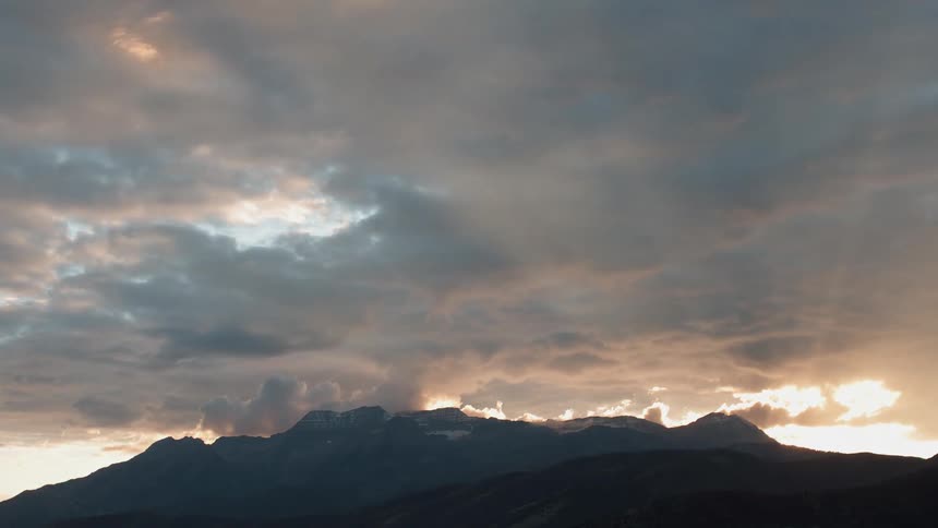 The sun is casting rays beyond mountains as it sets on a cloudy day